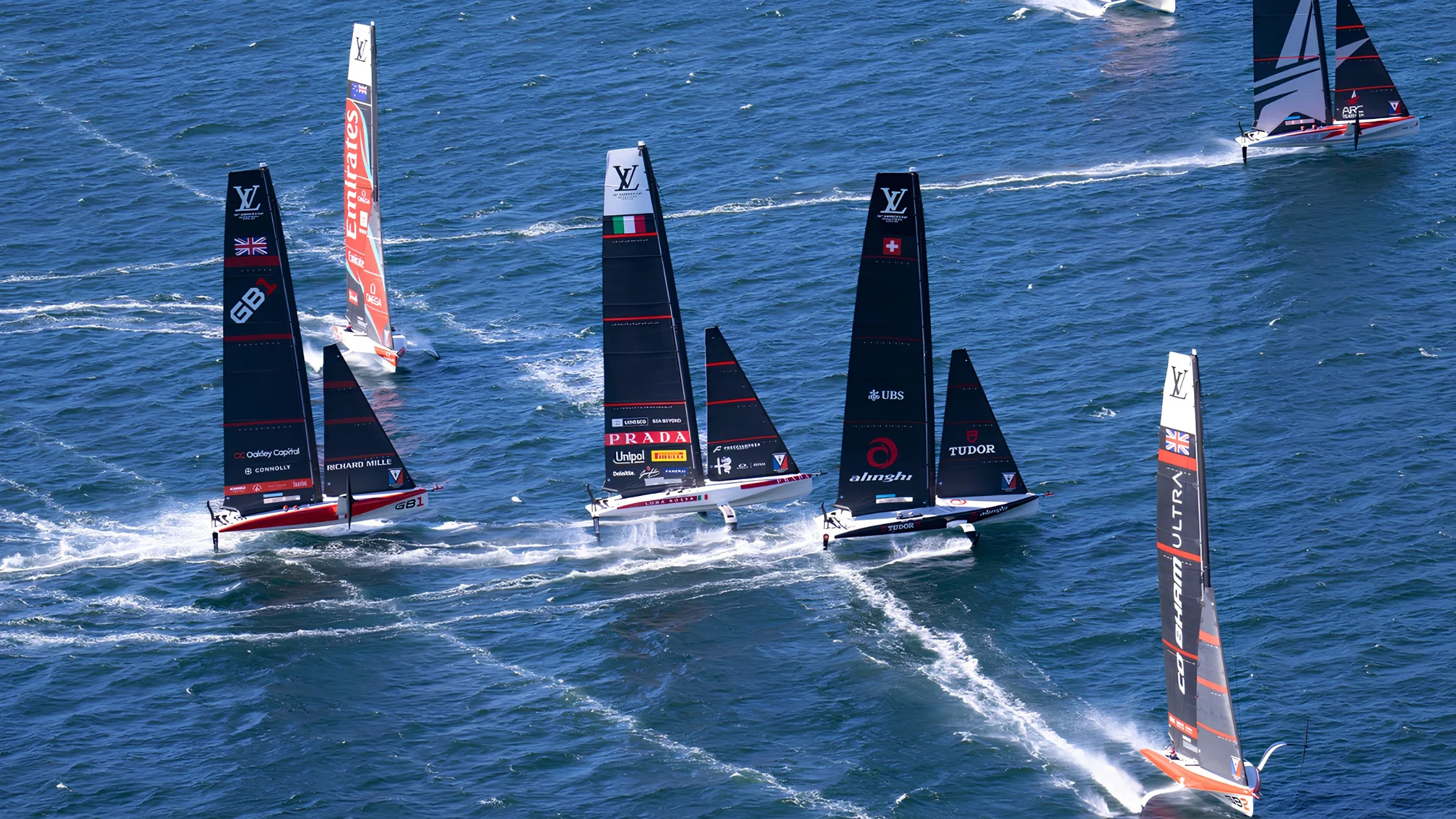 AC40 foiling yachts racing close together on blue water, seen from the air: GB1, Emirates Team New Zealand, Luna Rossa and Alinghi side by side, with GB2 and ARC Team USA at the edges of the fleet
